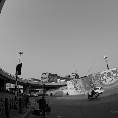 Bức ảnh chụp chân cầu Chương Dương tại một khu vực đô thị với các phương tiện giao thông như xe máy và ô tô cùng một người bán hàng đang di chuyển. Khung cảnh tạo cảm giác bình yên và nhịp sống thường nhật của thành phố.