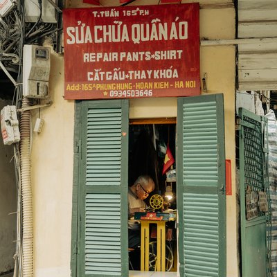Bức ảnh chụp một cửa hàng sửa chữa quần áo nhỏ tại Hà Nội với một người đàn ông lớn tuổi đang làm việc bên máy may. Không gian cửa hàng nhỏ gọn và ấm cúng, tạo cảm giác bình dị và gần gũi.