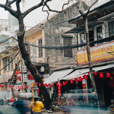 Bức ảnh chụp một góc phố Ngô Sĩ Liên với các toà nhà cũ, cây cối trụi lá và các xe máy đang di chuyển nhanh. Không khí nhộn nhịp và sống động của một con phố Hà Nội hiện lên rõ nét qua các hoạt động thường ngày và trang trí cờ hoa.