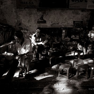 Bức ảnh đen trắng ghi lại cảnh một nhóm người ngồi nghỉ tại một quán nhỏ trên phố trong ánh nắng chói chang. Không khí trong ảnh mang lại cảm giác hoài niệm và chân thực về cuộc sống thường nhật ở thành phố.