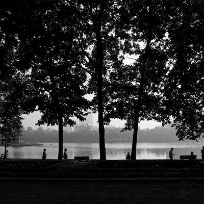 Bức ảnh đen trắng chụp Hồ Gươm với những cây xanh lớn và mặt nước hồ rộng. Không khí trong ảnh mang lại cảm giác yên bình và tĩnh lặng vào buổi sáng sớm.