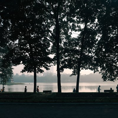Bức ảnh chụp cảnh Hồ Gươm với những cây xanh lớn và vài người đang đi dạo hoặc nghỉ ngơi bên bờ hồ vào buổi sáng. Không khí yên bình và tĩnh lặng tạo cảm giác thư thái cho người xem.