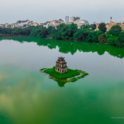 Hình ảnh chụp Hồ Hoàn Kiếm với mặt hồ rộng lớn, cây xanh bao quanh và kiến trúc cổ trên đảo nhỏ giữa hồ. Cảnh vật tạo cảm giác yên bình và thanh tĩnh giữa trung tâm thành phố nhộn nhịp.