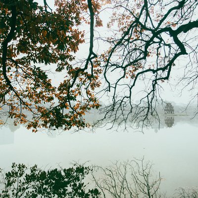 Bức ảnh chụp Hồ Gươm vào buổi sáng sớm với sương mù nhẹ bao phủ mặt hồ và những cành cây có lá vàng rơi. Không gian tạo cảm giác yên bình và tĩnh lặng, mang đến vẻ đẹp mơ màng của thủ đô Hà Nội.