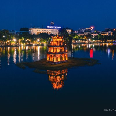 Hình ảnh Tháp Rùa nằm trên hồ Hoàn Kiếm vào ban đêm với ánh đèn vàng chiếu sáng và phản chiếu trên mặt nước. Cảnh vật mang lại cảm giác yên bình và tĩnh lặng giữa không gian đô thị sôi động.