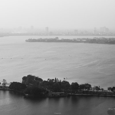 Bức ảnh chụp toàn cảnh Hồ Tây với cảnh thành phố Hà Nội mờ ảo trong sương mù phía xa. Không gian yên bình với mặt nước rộng lớn và cây xanh ven hồ tạo cảm giác thư thái.