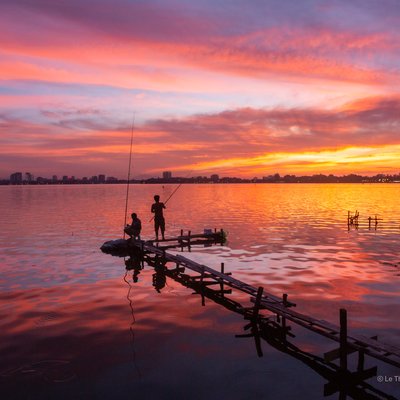 Bức ảnh chụp cảnh hoàng hôn trên hồ Tây với hai người đang câu cá trên một chiếc cầu gỗ nhỏ. Màu sắc ấm áp của bầu trời và mặt nước tạo nên không gian yên bình và thư thái.