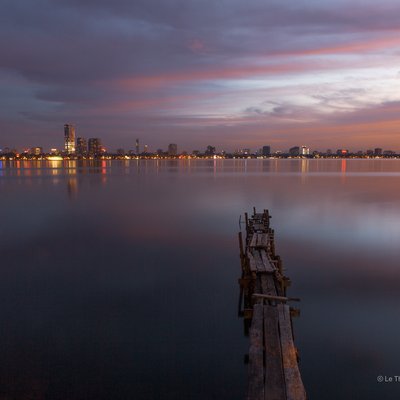 Bức ảnh chụp cảnh hoàng hôn trên Hồ Tây với cầu gỗ cũ dẫn ra mặt nước phẳng lặng, phía xa là ánh đèn của thành phố Hà Nội. Khung cảnh tạo cảm giác yên bình và tĩnh lặng trong khoảnh khắc chuyển giao giữa ngày và đêm.