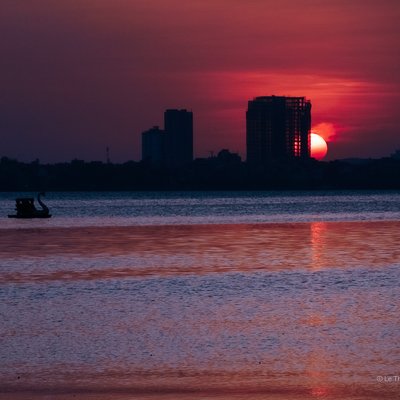 Bức ảnh chụp hoàng hôn trên hồ Tây với mặt trời đỏ rực gần chân trời và bóng các toà nhà cao tầng. Cảnh vật tạo cảm giác yên bình và tĩnh lặng khi ánh sáng dịu nhẹ phản chiếu trên mặt nước.