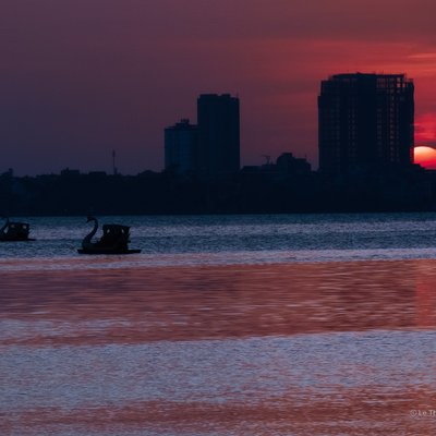 Bức ảnh chụp cảnh hoàng hôn trên hồ Tây với mặt trời đỏ rực gần chân trời và bóng các toà nhà cao tầng ở phía xa. Cảnh vật mang lại cảm giác yên bình và tĩnh lặng khi ánh sáng dịu dàng phản chiếu trên mặt nước.