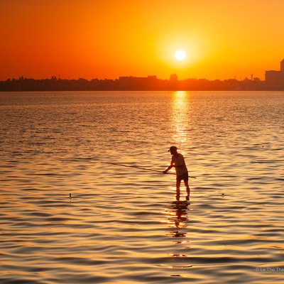 Ảnh chụp cảnh bình minh trên hồ Tây với một người đang câu cá trên mặt nước phẳng lặng. Không gian yên bình và ánh sáng vàng ấm áp tạo nên cảm giác thư thái và tĩnh lặng.