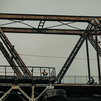 Bức ảnh chụp một cây cầu sắt cũ với vài người đang đứng và đi xe máy trên cầu. Không gian tạo cảm giác yên bình và tĩnh lặng trong một buổi chiều ở thành phố.