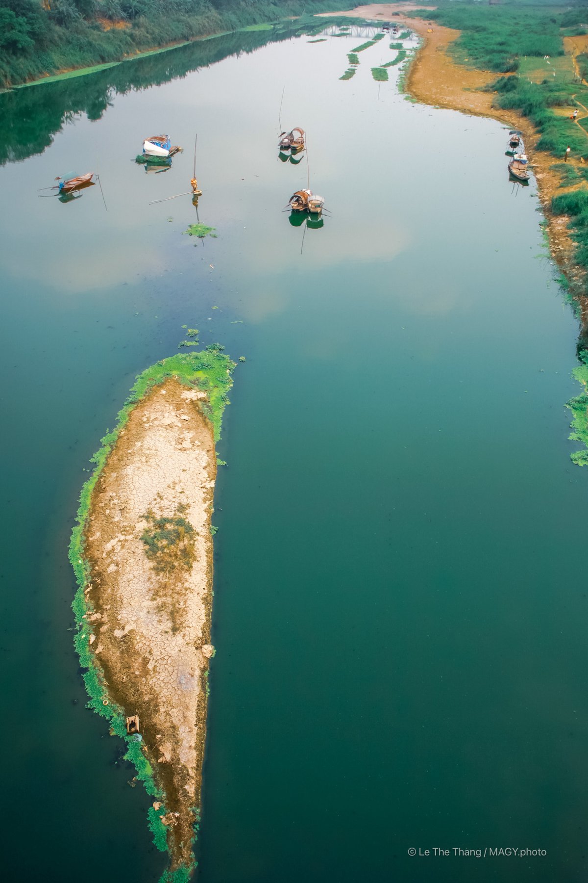 Bức ảnh chụp một đoạn sông với vài chiếc thuyền nhỏ neo đậu và một đảo nhỏ đất khô giữa dòng nước. Cảnh vật mang lại cảm giác yên bình và tĩnh lặng của vùng nông thôn vào ban ngày.