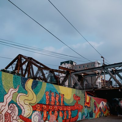 Bức ảnh chụp cầu Long Biên với bức tranh tường nhiều màu sắc bên dưới và một người đi bộ trên cầu. Cảnh vật tạo cảm giác yên bình trong một buổi chiều ở thành phố.