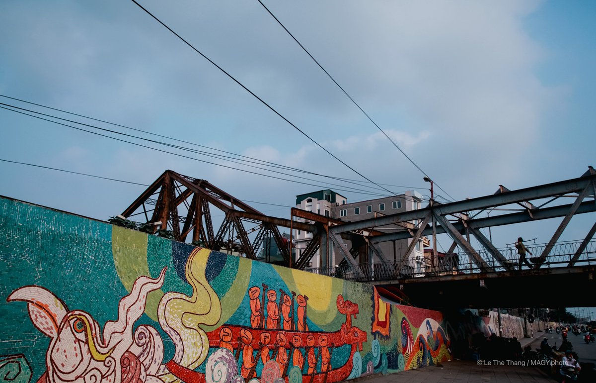 Bức ảnh chụp cầu Long Biên với bức tranh tường nhiều màu sắc bên dưới và một người đi bộ trên cầu. Cảnh vật tạo cảm giác yên bình trong một buổi chiều ở thành phố.
