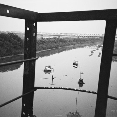 Bức ảnh chụp một con sông với vài chiếc thuyền nhỏ và cây cối ven bờ, nhìn qua khung kim loại của một công trình. Cảnh vật mang lại cảm giác yên bình và tĩnh lặng trong không gian rộng mở.