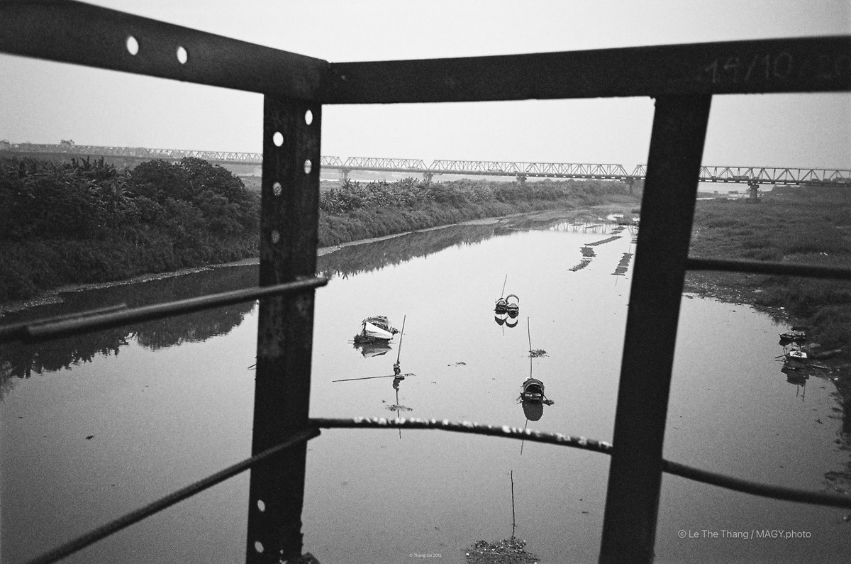 Bức ảnh chụp một con sông với vài chiếc thuyền nhỏ và cây cối ven bờ, nhìn qua khung kim loại của một công trình. Cảnh vật mang lại cảm giác yên bình và tĩnh lặng trong không gian rộng mở.