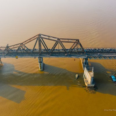 Hình ảnh chụp từ trên cao của cầu Long Biên bắc qua sông Hồng tại Hà Nội. Cảnh vật mang lại cảm giác yên bình với ánh sáng ban chiều dịu nhẹ và dòng sông chảy êm đềm.