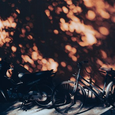 Bức ảnh chụp hai chiếc xe, một xe máy và một xe đạp, đậu bên cạnh một bức tường với ánh nắng chiếu qua tạo ra các bóng râm trên tường. Cảnh vật mang lại cảm giác yên bình và tĩnh lặng trong một buổi chiều tại thành phố.