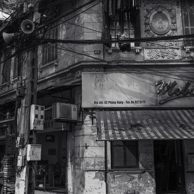 Bức ảnh chụp một ngôi nhà cũ trên phố Phùng Hưng với nhiều chi tiết như dây điện chằng chịt, ghế nhựa và bảng hiệu đã phai màu. Hình ảnh gợi lên cảm giác hoài niệm về một Hà Nội xưa cũ với không gian sống giản dị và thân thuộc.