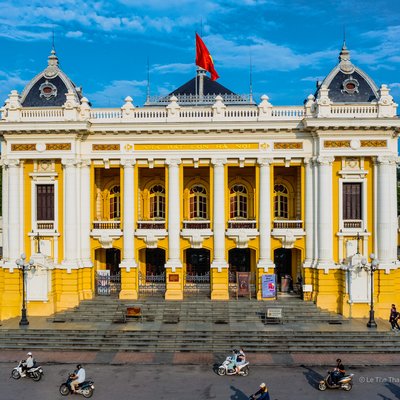 Hình ảnh nhà hát lớn Hà Nội với kiến trúc cổ điển nổi bật dưới ánh sáng ban ngày. Cảnh vật xung quanh có người đi xe máy tạo nên không khí sống động của thành phố thủ đô.