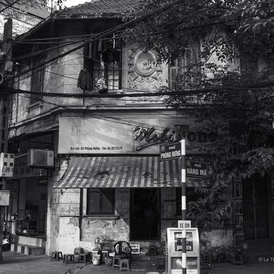 Bức ảnh chụp một góc phố cổ Hà Nội với quán cà phê nhỏ bên đường, các chi tiết như bảng hiệu, ghế nhựa và xe máy tạo nên không khí đặc trưng. Cảnh vật mang lại cảm giác hoài niệm về một Hà Nội truyền thống yên bình và giản dị.