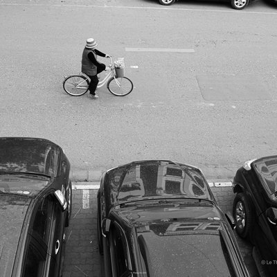 Bức ảnh chụp một người đang đi xe đạp trên đường phố có nhiều ô tô đỗ bên cạnh. Cảnh vật tạo cảm giác bình yên và giản dị trong cuộc sống đô thị.