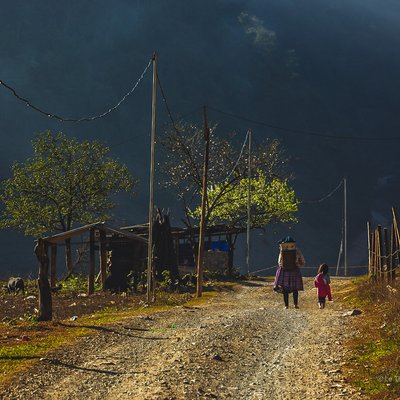 Bức ảnh chụp một con đường đất ở vùng nông thôn Mộc Châu với hai người đang đi bộ, xung quanh có cây xanh và nhà đơn sơ. Khung cảnh mang lại cảm giác yên bình và giản dị của cuộc sống thường ngày nơi đây.