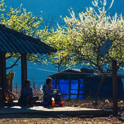 Bức ảnh chụp cảnh hai người phụ nữ ngồi nghỉ trên hiên nhà ở vùng nông thôn Mộc Châu, dưới ánh sáng mềm mại của buổi chiều. Cảnh vật mang lại cảm giác bình yên và truyền thống, thể hiện cuộc sống thường nhật giản dị.