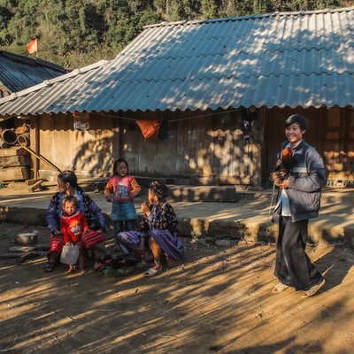 Bức ảnh chụp một nhóm người trong bản làng Mộc Châu, Sơn La vào buổi chiều với ánh nắng nhẹ chiếu qua. Cảnh vật mang lại cảm giác yên bình và ấm áp của cuộc sống vùng cao truyền thống.