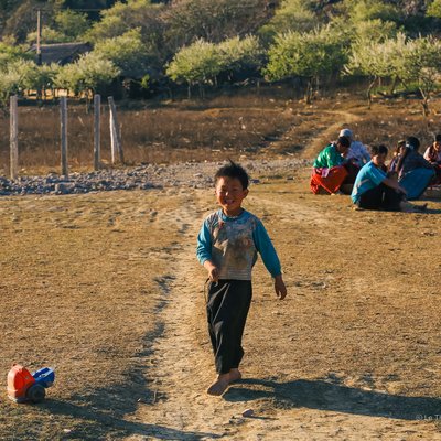 Bức ảnh chụp một đứa trẻ đang chơi đùa trên con đường đất trong một khu vực nông thôn với nhóm người ngồi phía sau. Cảnh vật tạo cảm giác bình yên và vui vẻ, thể hiện cuộc sống giản dị của trẻ em vùng cao.