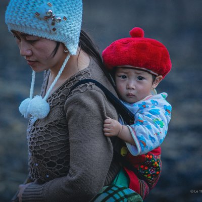 Bức ảnh chụp một người phụ nữ mang theo đứa trẻ trên lưng trong trang phục truyền thống ở vùng cao Mộc Châu. Cảnh vật tạo cảm giác yên bình và gợi lên tình cảm gia đình ấm áp.