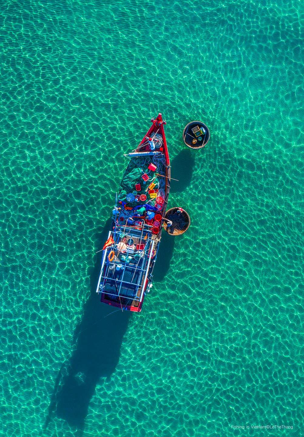 Bức ảnh chụp toàn cảnh trên không một chiếc thuyền đánh cá truyền thống trên vùng biển trong xanh. Khung cảnh tạo cảm giác bình yên và tĩnh lặng với ánh sáng tự nhiên dịu nhẹ chiếu xuống mặt nước.