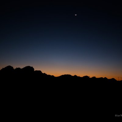 Bức ảnh chụp cảnh hoàng hôn tại Mộc Châu với bầu trời xanh thẫm và trăng lưỡi liềm trên cao, phía dưới là các đồi núi đen bóng. Cảnh vật mang lại cảm giác yên bình và tĩnh lặng, tạo nên một khung cảnh thiên nhiên tuyệt đẹp.