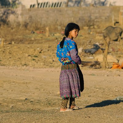 Bức ảnh chụp một bé gái mặc trang phục thổ cẩm truyền thống đứng ngoài trời ở vùng nông thôn Mộc Châu. Cảnh vật xung quanh tạo cảm giác yên bình và tĩnh lặng vào buổi chiều.