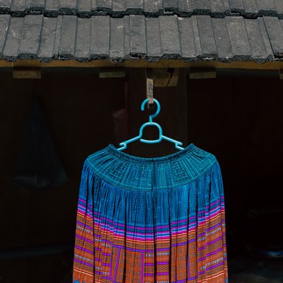 Chiếc váy thổ cẩm với hoạ tiết đặc trưng của Mộc Châu, Sơn La được treo trên móc nhựa màu xanh dưới mái nhà. Hình ảnh toát lên vẻ đẹp truyền thống và sự tinh tế trong văn hoá địa phương.