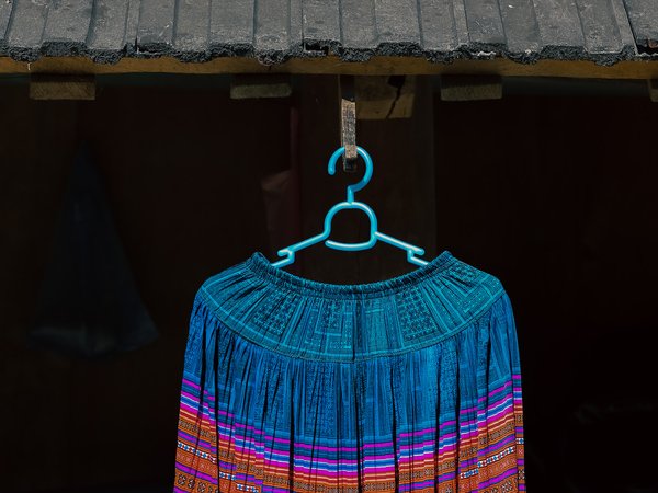 Chiếc váy thổ cẩm với hoạ tiết đặc trưng của Mộc Châu, Sơn La được treo trên móc nhựa màu xanh dưới mái nhà. Hình ảnh toát lên vẻ đẹp truyền thống và sự tinh tế trong văn hoá địa phương.