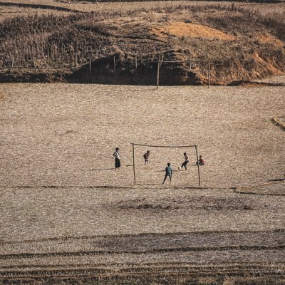 Bức ảnh chụp một nhóm trẻ em đang chơi bóng trên một cánh đồng rộng lớn ở vùng nông thôn. Khung cảnh tạo cảm giác yên bình và giản dị của cuộc sống thường nhật nơi đây.