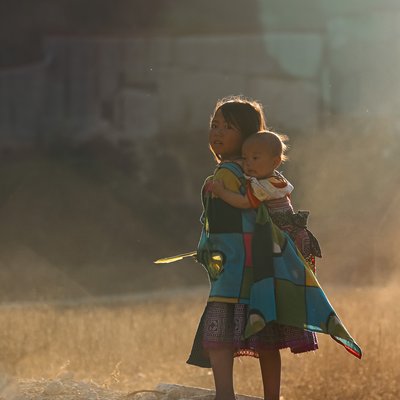 Bức ảnh chụp hai trẻ em ở Mộc Châu, một bé gái đang cõng em nhỏ trên lưng trong ánh sáng chiều dịu nhẹ. Cảnh vật tạo cảm giác bình yên và truyền thống của cuộc sống nông thôn.