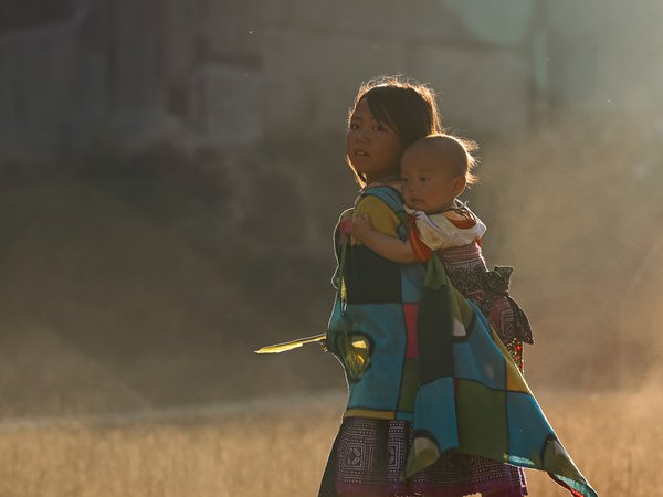 Bức ảnh chụp hai trẻ em ở Mộc Châu, một bé gái đang cõng em nhỏ trên lưng trong ánh sáng chiều dịu nhẹ. Cảnh vật tạo cảm giác bình yên và truyền thống của cuộc sống nông thôn.