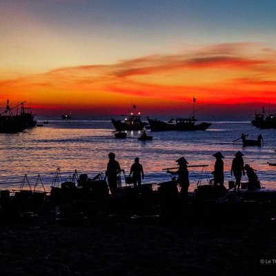 Bức ảnh chụp cảnh chợ cá Tam Tiến bên bờ biển Quảng Nam vào lúc hoàng hôn với những chiếc thuyền đánh cá và người dân đang làm việc. Khung cảnh tạo cảm giác yên bình và truyền tải nét đẹp của cuộc sống thường nhật miền Trung Việt Nam.
