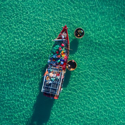 Hình ảnh chụp từ trên cao cho thấy một chiếc tàu cá và hai thuyền thúng trên mặt nước biển trong xanh tại chợ cá Tam Tiến, Quảng Nam. Cảnh vật tạo cảm giác yên bình và truyền tải nét đẹp truyền thống của cuộc sống ven biển miền Trung.