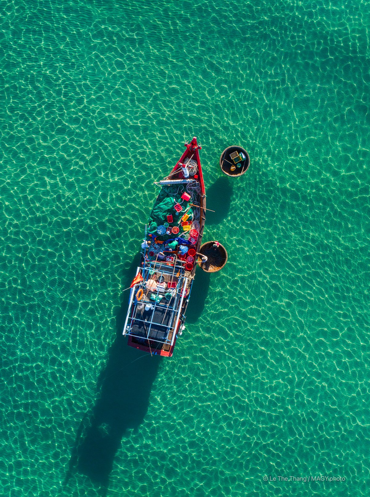 Hình ảnh chụp từ trên cao cho thấy một chiếc tàu cá và hai thuyền thúng trên mặt nước biển trong xanh tại chợ cá Tam Tiến, Quảng Nam. Cảnh vật tạo cảm giác yên bình và truyền tải nét đẹp truyền thống của cuộc sống ven biển miền Trung.