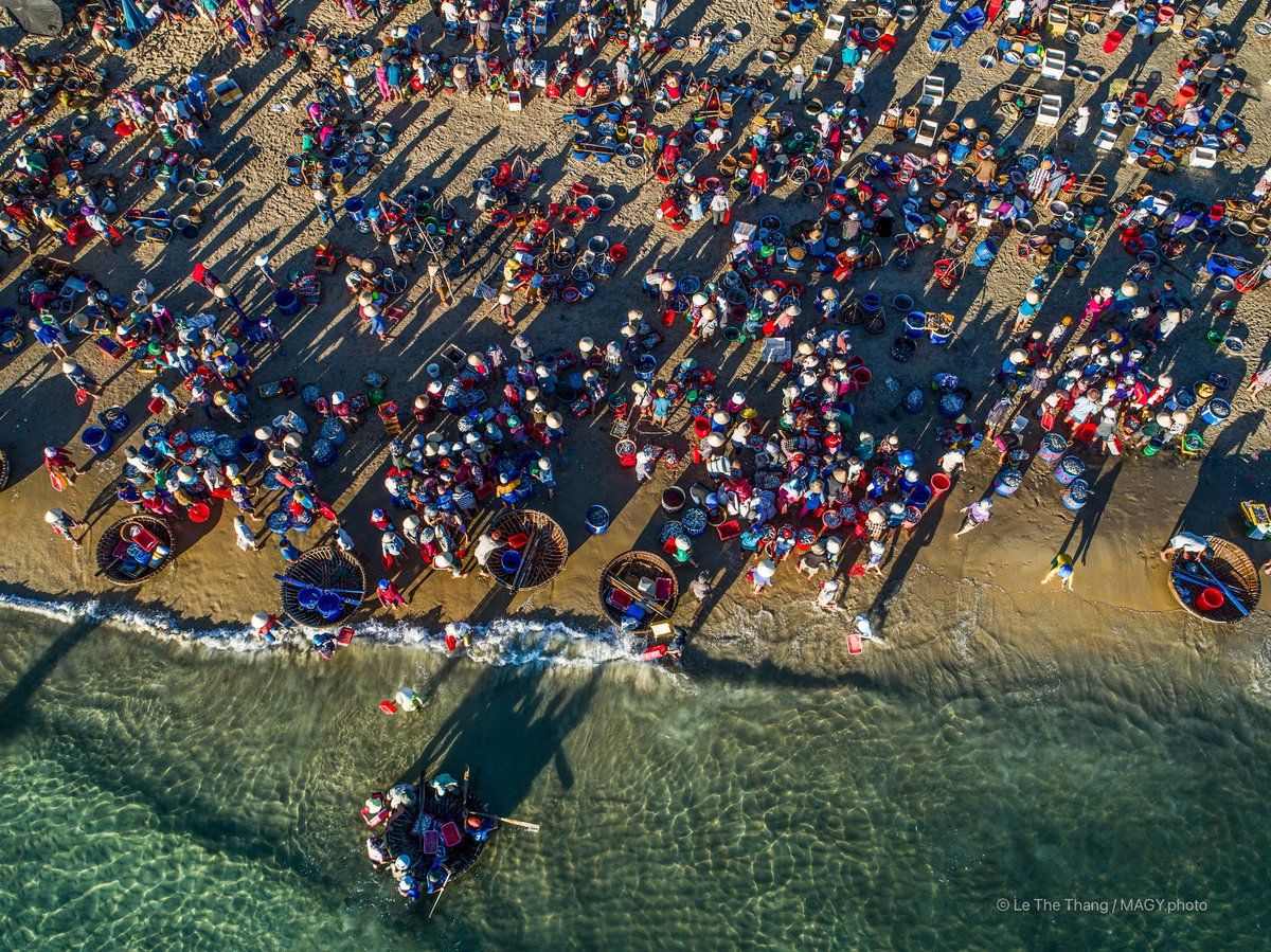 Ảnh chụp từ trên cao cho thấy cảnh nhộn nhịp của chợ cá Tam Tiến bên bờ biển với nhiều người và thuyền thúng. Không khí sôi động và đầy màu sắc thể hiện cuộc sống truyền thống của ngư dân miền Trung.