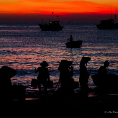 Hình ảnh chợ cá Tam Tiến tại Quảng Nam vào lúc hoàng hôn, với các bóng người và thuyền trên biển. Cảnh vật tạo cảm giác yên bình và truyền tải nét đẹp của cuộc sống ngư dân miền Trung.