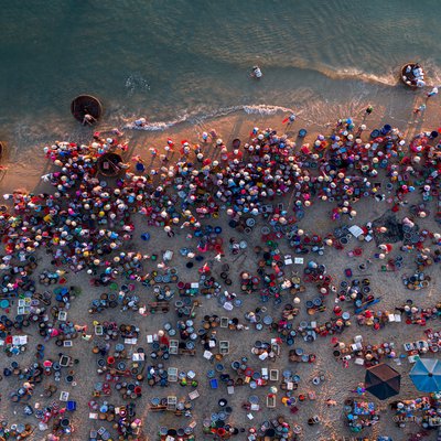 Hình ảnh chụp từ trên cao cho thấy một khu chợ cá đông đúc bên bờ biển với nhiều người và thuyền thúng. Không khí sôi động và nhộn nhịp của chợ cá truyền thống miền Trung được thể hiện rõ qua màu sắc và hoạt động của người dân.