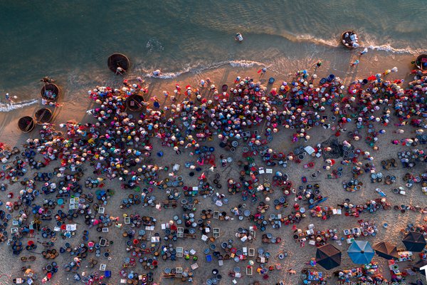 Hình ảnh chụp từ trên cao cho thấy một khu chợ cá đông đúc bên bờ biển với nhiều người và thuyền thúng. Không khí sôi động và nhộn nhịp của chợ cá truyền thống miền Trung được thể hiện rõ qua màu sắc và hoạt động của người dân.
