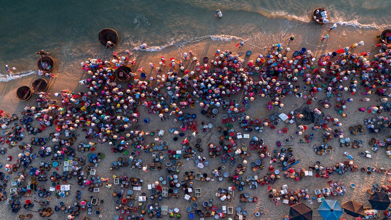 Hình ảnh chụp từ trên cao cho thấy một khu chợ cá đông đúc bên bờ biển với nhiều người và thuyền thúng. Không khí sôi động và nhộn nhịp của chợ cá truyền thống miền Trung được thể hiện rõ qua màu sắc và hoạt động của người dân.