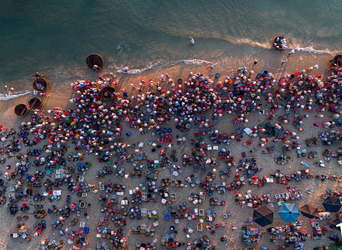 Hình ảnh chụp từ trên cao cho thấy một khu chợ cá đông đúc bên bờ biển với nhiều người và thuyền thúng. Không khí sôi động và nhộn nhịp của chợ cá truyền thống miền Trung được thể hiện rõ qua màu sắc và hoạt động của người dân.