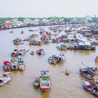 Hình ảnh chợ nổi Cái Răng với nhiều thuyền buôn tấp nập trên sông, các mái nhà và cây xanh hai bên bờ. Toàn cảnh tạo cảm giác sống động và nhộn nhịp của một khu chợ truyền thống trên sông.